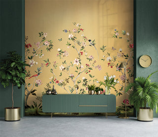 Crown Surface Solutions Daisy Bennett wallpaper in a room setting featuring a chinoiserie mural of floral branches and birds on a warm yellow background, styled with a green console, potted plants, and gold floor lamp.
