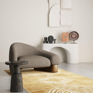 Modern armchair in textured brown JF Fabrics Lennon collection upholstery. Features a curved silhouette on a wood base, set in an organic interior with a golden geometric rug, dark side table, and a white arched console with minimalist sculptures.