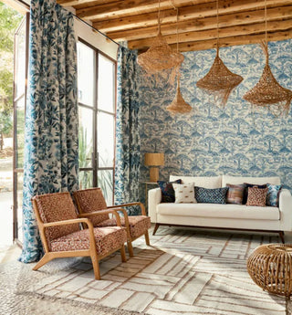 Fabric collection styled in a living room featuring blue and white tropical print drapes paired with a matching blue scenic toile wallpaper backdrop, complementary upholstered armchairs, a cream sofa, and woven pendant lights.