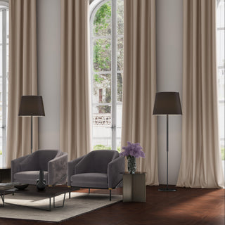 Floor-to-ceiling drapes from the JF Fabrics Pixie collection in Mushroom beige frame large arched windows. Two charcoal velvet armchairs sit on a beige rug with dark wood furniture and black floor lamps, accented by a bouquet of purple flowers.