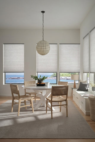 A light coastal dining room with a white table and cane chairs. Grey textured cellular shades are on the large windows, showing an extended view of the rocky ocean shoreline below. A white storage bench with pillows is on the right, and a large grid-patterned globe pendant light hangs above the table.