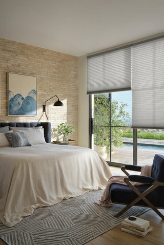 A modern bedroom with a brick wall, art, and a large bed. On the right, large windows with grey cellular blinds overlook an outdoor deck with a pool. A navy lounge chair sits on a geometric rug.