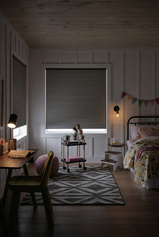 A cozy girl's bedroom with vertical panelling and two windows with closed grey cellular shades. A desk with a lamp and an open notebook sits on the left, and a bed with a colourful quilt sits on the right. A plant stand, pouf, and step stool are between them on a geometric rug. Small colourful bunting and a warm sconce add charm. A unique light strip glows at the base of the blinds.