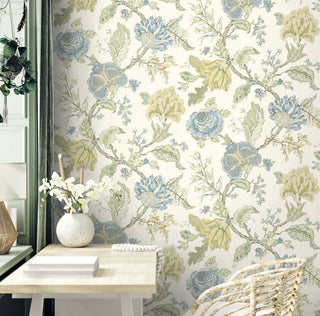 A light wooden desk sits against Crown’s Provence Aries wallpaper, featuring large hand‑drawn florals and leafy branches in soft blue, green, and beige on a cream background. A white vase with flowers, stacked books, and a beaded accent rest on the desk. A green wall panel, hanging plant, and woven chair add natural texture, creating a calm, botanical-inspired space.