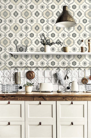 A cozy kitchen with cream cabinetry and a wooden countertop sits beneath Crown’s Provence Avignon wallpaper, featuring a soft gray and cream geometric floral motif with a vintage French feel. A white embossed tile backsplash, floating shelf with vases and jars, wire baskets, a toaster, and a small plant add rustic charm. A metal pendant light hangs above, illuminating the warm, lived‑in space.