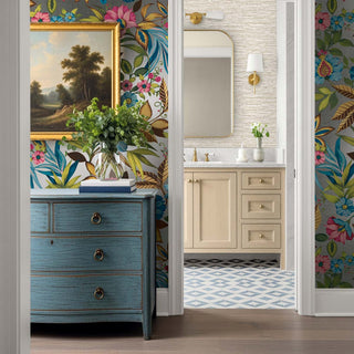 A styled interior scene showing a bathroom vanity area with colorful floral wallpaper, a blue chest of drawers, a framed landscape painting, and a mirror. The likely product being highlighted is the wallpaper, shown in a real room environment rather than as an isolated swatch.
