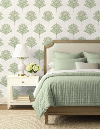 A styled bedroom scene featuring a bed with sage green bedding, a white nightstand with a lamp and flowers, and wallpaper with a repeating green palm leaf pattern on a white background.