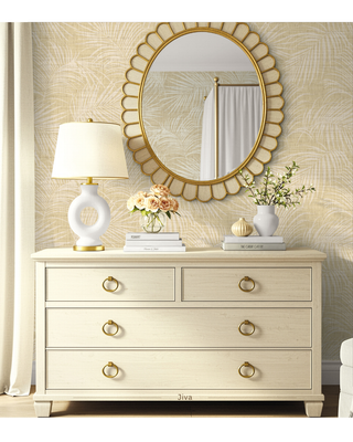 A light cream-colored wooden dresser with brass ring pulls, styled with a white ceramic table lamp, books, and flowers, beneath a scalloped gold-framed mirror against a patterned wallpaper background.