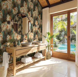 A modern bathroom vanity with two vessel sinks, a round mirror, and a light wood console table, set against a tropical palm-patterned wall. Large glass doors look out to a resort-style pool area with palm trees.