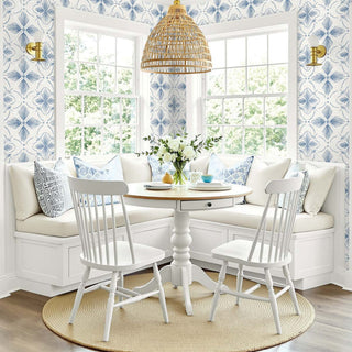 A bright coastal-style dining nook with a white round pedestal table, two white spindle-back chairs, a woven pendant light, and a built-in white bench with blue and white patterned pillows. The room features blue and white patterned wallpaper, gold wall sconces, and a round jute rug on a wood floor.