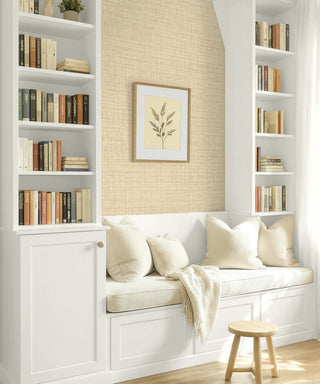 A cozy built-in white bookcase and bench with cream cushions and a throw blanket, set against a textured beige wall with a framed botanical print. White built-in shelves on both sides are filled with books, and a small wooden stool sits in the foreground.