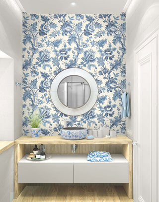A bathroom setting featuring the Cabris wallpaper from the Crown Provence collection on a focal wall. The wallpaper showcases an elegant blue and cream Toile de Jouy floral and botanical design. Centered on the wall is a round white-framed mirror hanging above a light-wood vanity counter. A blue and white patterned ceramic vessel sink, a matching potted plant, and bathroom accessories sit on the counter. Below, a floating white cabinet provides storage, creating a bright, elegant, and classic space.