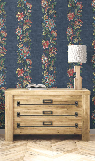 A room setting featuring the Callas wallpaper from the Crown Provence collection on a full wall. The wallpaper showcases vertical trails of rich, folk-inspired floral motifs in red, pink, and green against a textured denim blue background. In front of the wall, a rustic, light-wood dresser with industrial black metal drawer pulls stands on a light chevron-patterned wood floor. A whimsical, textured white lamp on a blocky wood base and an open book rest on top of the chest, creating a cozy vibe.