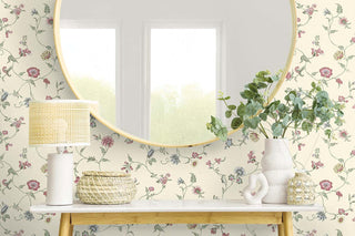 A console table with a white marble top stands against Crown’s Provence collection wallpaper in the Cassis pattern, featuring delicate trailing florals in soft pinks, purples, and greens on a warm cream background. The table holds a woven basket, sculptural ceramic piece, beige vase with greenery, small container, and a white lamp with a textured shade. Above it hangs a round gold-framed mirror reflecting bright windows.