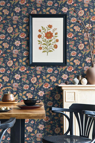 A room setting featuring the Castellane wallpaper from the Crown Provence collection on a full wall. The design showcases sprawling botanical floral trails with detailed orange, cream, and green blossoms on a dark charcoal-blue background. A black-framed art print displaying a matching floral sprig hangs prominently. In the foreground, a rustic wooden table holds a tea set, flanked by a blue spindle-back chair. A light cream mantelpiece is visible to the right, creating a warm, cozy atmosphere.