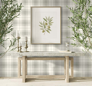 A bright entryway setting featuring the Gordes wallpaper from the Crown Provence collection on a full wall. The wallpaper displays a classic country cottage plaid design with soft sage-grey and cream checks accented by thin geometric grid lines over a linen-textured background. A framed art print of an olive branch hangs centered above a rustic wooden console table with a thick stone countertop. Tall potted olive trees flank the table, creating a serene, natural, and welcoming aesthetic.