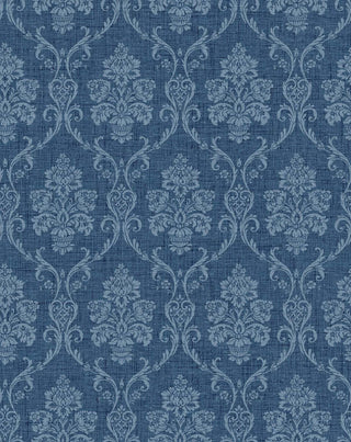 A close-up view of the Istres wallpaper swatch from the Crown Provence collection. The pattern features an ornate, classic damask motif showcasing elegant floral bouquets in stylized urns, enclosed by delicate scrolling acanthus leaves. The intricate design is rendered in a blue tone that sits subtly against a warm, denim blue background. The backdrop is enhanced with a fine, linen-textured faux weave, giving the traditional pattern a tactile, fabric-like depth and a serene, sophisticated feel.