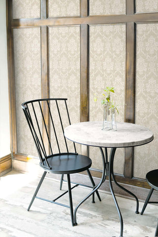 A bistro setting featuring the Istres wallpaper from the Crown Provence collection inlaid within dark wood wall paneling. The wallpaper displays an elegant cream damask floral motif on a soft beige, linen-textured background. In the foreground, a modern black spindle-back chair sits next to a small round cafe table with a light stone-mosaic top. A collection of slender glass vases with delicate green plant sprigs rests on the table, creating a bright, sophisticated, and contemporary cafe atmosphere.