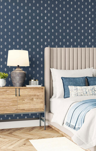 A bedroom setting featuring the Sault wallpaper from the Crown Provence collection on the wall behind a bed. The wallpaper displays a small, repeating white damask-inspired motif on a textured navy blue background. A bed with a high, channel-tufted beige headboard is dressed in white linens, denim blue accent pillows, and a light blue throw blanket. To the left, a natural wood nightstand holds a dark, textured ceramic lamp with a glowing white shade, adding a warm, sophisticated ambiance.