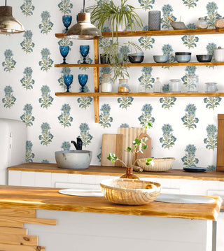A room setting featuring the Sellans wallpaper from the Crown Provence collection on a kitchen wall behind open wooden shelving. The wallpaper showcases an elegant, repeating botanical block-print floral motif in soft French blue and sage green on a crisp cream background. The wooden shelves display blue glassware, ceramic bowls, and jars. Below, a natural wood countertop holds a stone sink basins, cutting boards, and woven baskets, creating a warm, rustic, and inviting cottage aesthetic.