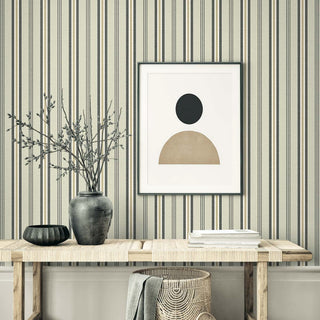 A minimalist console table sits against Crown’s Provence Toulon wallpaper, featuring refined vertical stripes in soft beige, gray, and cream with a subtly textured finish. A dark ceramic vase with branches, a black bowl, and stacked books rest on the wooden table. Below, a woven basket with a draped gray cloth adds warmth. Above, a framed abstract print completes the calm, modern composition.
