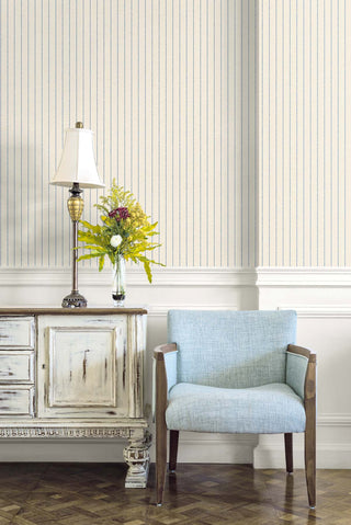 A room setting featuring the Tourves wallpaper from the Crown Provence collection on the upper half of a wall, above white wainscoting. The wallpaper has thin, vertical blue ticking stripes on a cream background. In the foreground, a light blue upholstered armchair with wooden arms sits on a parquet floor next to a distressed white wooden sideboard. On the sideboard, a traditional table lamp with a white shade and a glass vase with a yellow and green floral arrangement add a cozy charm.