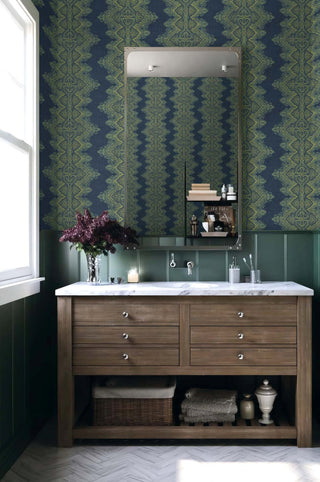A bathroom with a wooden vanity and marble countertop, topped with a vase of dark flowers and a lit candle. A large mirror with a small shelf hangs above. Behind it, the Valensole wallpaper from Crown’s Provence collection shows a blue and green zigzag motif above dark green paneling. A window brightens the space, which includes herringbone flooring and neatly arranged towels and décor.