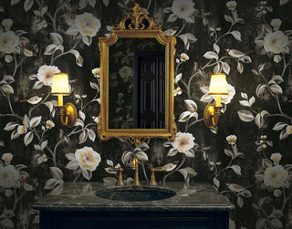 Moody powder room featuring Crown's Chinoiserie collection wallpaper in the Arang Floral Trail pattern. The dramatic walls display a textured charcoal-black background adorned with climbing vines, lush cream and blush camellia blossoms, and small perched birds. Centered is an ornate, heavily detailed gold mirror hanging above a dark gray veined marble vanity with a classic brass faucet. Two matching vintage-style gold wall sconces with pleated cream shades flank the mirror, casting a warm glow.