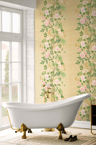 Elegant bathroom featuring Crown's Chinoiserie collection wallpaper in the Jangmi Trail pattern, displaying vertical climbing branches with pink peonies and yellow blossoms on a textured gold background. In the foreground, a white clawfoot bathtub with ornate brass feet sits on a patterned rug next to a pair of leopard print flats. A classic gold floor-mount faucet stands behind the tub. To the left, a large grid window and a white painted brick wall fill the space with natural light.