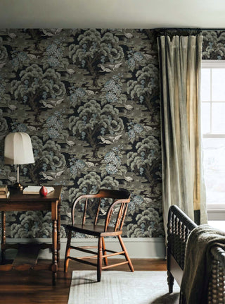Vintage bedroom setting featuring Crown's Chinoiserie collection wallpaper in the Hanae Trees pattern. The wall showcases a stylized, artistic tree trunk climbs vertically, showcasing painterly branches in soft charcoal gray. The tree is densely packed with large, rounded, overlapping flower heads or foliage clusters in contrast. Against the wall stands a contemporary console table with a black frame and light wood drawer. On top, a matching wooden organizer holding a notepad rests under a soft glowing lamp