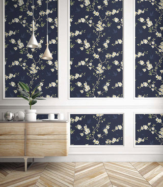 Elegant room setting featuring Crown's Chinoiserie collection wallpaper in the Hanami Trail pattern. The design is displayed inside decorative white wall molding panels against a dark navy blue background. Slender, winding branches climb upward, heavily dotted with delicate clusters of white and soft cream plum blossoms with golden centers. Faint silhouette shapes of birds and butterflies glide through the background. Below, a light wood console table stands on a light-colored chevron wood floor.