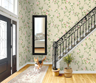 Bright entryway featuring Crown's Chinoiserie collection wallpaper in the Jangmi Trail pattern, which displays delicate climbing vines, pink and white roses, and small birds on a cream background. A black ornate wrought-iron staircase railing lines carpeted stairs on the right, next to a small potted plant. A long black-framed mirror hangs centrally. On the light hardwood floor, a tan and black dog joyfully runs across a vintage-style runner rug near a dark gray double front door.