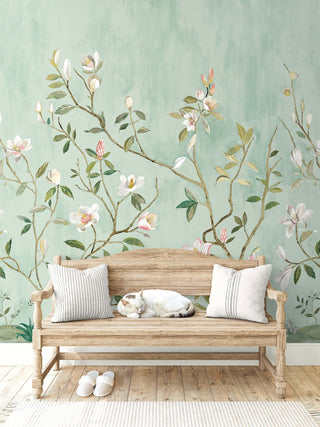 A serene entryway with a wooden bench holding two light cushions and a resting cat, set on a warm wood floor with white slippers placed neatly in front. Behind the bench, the Kaiyu Dogwood Mural from Crown’s Chinoiserie collection shows soft green tones with delicate branches, white and pink blossoms, and leafy details, creating a calm, nature‑inspired backdrop.