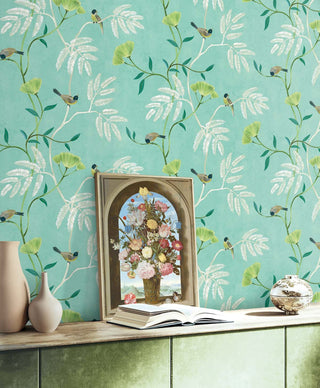 Bright room setting featuring Crown's Chinoiserie collection wallpaper in the Kohana Branch pattern. The wall showcases a textured, light teal background adorned with an elegant botanical trail of silver-gray and gold branches. The branches feature stylized, fan-shaped chartreuse blossoms, delicate fern-like pinnate leaves, and small, detailed perched songbirds. Against the wall is a long sideboard holding an arched, framed floral still life painting, an open book, and decorative ceramic vases.