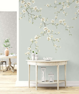 An interior scene with a light wooden console table decorated with white vases, small green plants, a coral‑shaped ornament, and two lanterns. Behind it, the Miyabi Bloom Mural from Crown’s Chinoiserie collection shows soft magnolia branches with white blossoms on a pale background. The space feels calm, airy, and elegantly styled, with a glimpse into an adjoining room.