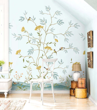 A cozy corner with a white wooden chair beside a stacked set of round wooden boxes topped with a woven‑shade lamp. A cushioned bench and a small plant sit nearby on warm wood flooring. Behind them, the Mokusel Branch Mural from Crown’s Chinoiserie collection shows golden branches with green and blue leaves, yellow flowers, and small birds, creating a serene, artistic backdrop