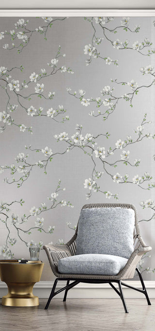 Serene room setting featuring Crown's Chinoiserie collection wallpaper in the Moonlace Magnolia pattern. The wallpaper showcases a textured, silvery-gray faux-grasscloth background with delicate, sprawling branches covered in elegant white magnolia blossoms and green buds. In the foreground, a stylish woven wicker armchair with a textured blue-gray cushion sits on a stone floor. A small, sculptural metallic gold side table holds a glass tumbler next to the chair, completing the modern look.