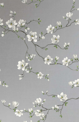 Close-up swatch of Crown's Chinoiserie collection wallpaper in the Moonlace Magnolia pattern. The elegant design features a light grey-silver background with a faint, block-grid texture overlaid by subtle, swirling line details. Graceful, dark grey branches twist across the canvas, bearing large, delicate white magnolia blossoms with white and small green leaves and buds. The botanical motif offers a luxurious, serene, and sophisticated aesthetic