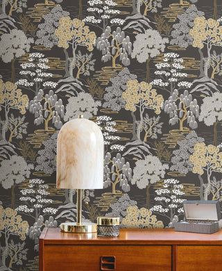 Atmospheric room setting featuring Crown's Chinoiserie collection wallpaper in the Mori Trees pattern. The wall is adorned with a detailed, woodblock-style forest illustration against a charcoal-black background, showcasing stylized trees in metallic gold, gray, and white. In the foreground, an elegant dome-shaped marble table lamp with a brass base sits on a mid-century wooden dresser. Next to the lamp are a small glass jar with a gold lid and an open gray textured jewelry box