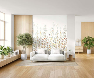 A modern living room with a white sofa on a beige rug, surrounded by potted green plants and light wood accents. Behind the sofa, the Sakura Bloom Mural from Crown’s Chinoiserie collection displays delicate branches with multicolored blossoms on a soft background. The space feels airy, warm, and nature‑inspired with clean, minimalist styling.