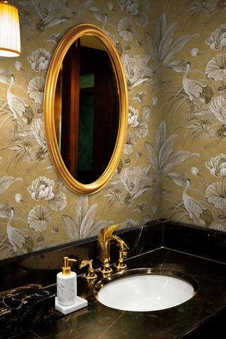 Luxurious powder room featuring Crown's Chinoiserie collection wallpaper in the Tsuru Bird pattern. The walls display an opulent scene of cranes, blooming lotus flowers, and tropical fronds in sepia tones on a distressed metallic gold background. A large oval mirror with a thick gold frame hangs above a black marble vanity with gold veining. The under-mount white sink is fitted with an ornate, sculptural gold faucet. A marble soap dispenser and black tissue box sit nearby.