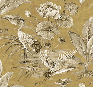 Close-up swatch of Crown's Chinoiserie collection wallpaper in the Tsuru Bird pattern. The detailed scenic design features a warm, antiqued metallic gold background with a subtle, distressed plaster patina. Beautifully rendered in classic sepia tones are elegant cranes or storks amidst a serene wetland; one stands tall with its beak open, while another glides in mid-flight over the water. Large blooming lotus flowers, rounded lily pads, seed pods, and lush tropical fronds complete the opulent landscape.