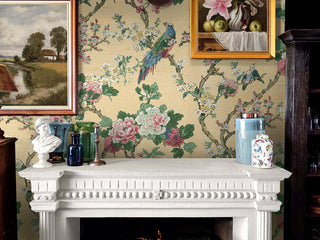 Elegant living room fireplace setting featuring Crown's Chinoiserie collection wallpaper in the Ume Bird Trail pattern. The wall showcases a textured, golden-yellow grasscloth background decorated with gnarled branches, vibrant pink peonies, and white blossoms. A colorful, long-tailed pheasant sits on a branch above a white ornate mantlepiece. Framed oil paintings hang above the fireplace, while a white bust, colored glass vases, and patterned ceramic jars decorate the mantel.