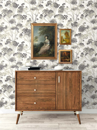 Sophisticated room setting featuring Crown's Chinoiserie collection wallpaper in the Yulin Trees pattern. The wall is covered in a distressed off-white design featuring taupe branches topped with fan-shaped clusters of charcoal-gray needles. Centered against the wall is a mid-century modern wooden credenza with three drawers and a side cabinet. Three ornate gold-framed vintage oil paintings hang above it. Stacked books and a white decorative bowl rest on top, all set on light wood floors