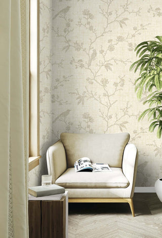 Bright reading nook featuring Crown's Chinoiserie collection wallpaper in the Yuye Silhouette pattern. The wallpaper showcases a textured, warm beige linen-like background decorated with soft gray branching trails, delicate blossoms, and perched birds. A plush cream armchair holds an open magazine next to a sunlit window with a draped curtain. A rustic wooden block side table with stacked books sits on elegant light chevron hardwood floors, completing the serene space