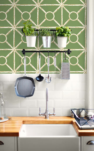 Modern kitchen backsplash area featuring Crown's Chinoiserie collection wallpaper in the Zhen Bamboo pattern. The wall features a vibrant sage green background overlaid with a realistic cream and light beige faux-bamboo trellis pattern forming a geometric square, diamond, and ring grid. Below the wallpaper is a classic white subway tile backsplash, a wooden countertop with a white apron sink, and a black rail hanging pots, kitchen utensils, and potted fresh herbs.